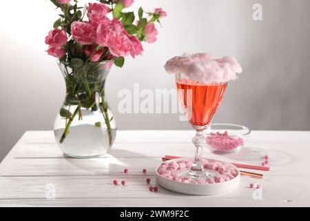 Cocktail de barbe à papa en verre, guimauves, vase avec des roses roses et des pailles roses sur une table en bois blanc sur fond gris Banque D'Images