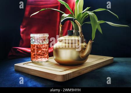 Une cruche en aluminium doré avec un verre de thé à côté, une plante sur une planche de bambou et un tissu de satin rouge Banque D'Images