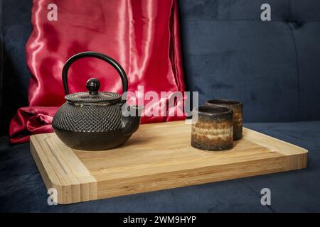 Un pichet en fer forgé noir de style asiatique avec quelques verres à thé à côté sur une planche de bambou avec un chiffon de satin rouge Banque D'Images