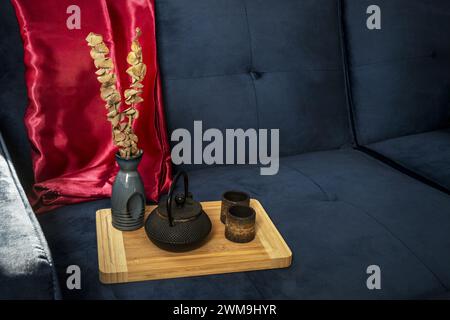 Une cruche en fer forgé noir de style asiatique avec quelques verres à thé à côté sur une planche de bambou avec un tissu de satin rouge sur un canapé en velours bleu Banque D'Images