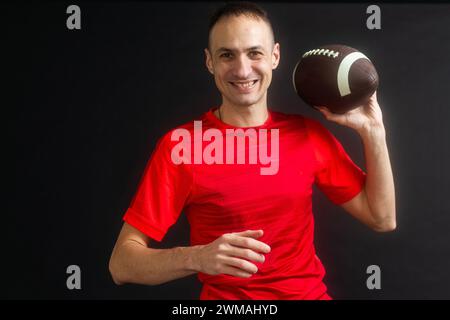 Jeune homme vêtu de vêtements de rue décontractés tient un ballon de football, prêt à lancer une passe, isolé sur fond noir Banque D'Images