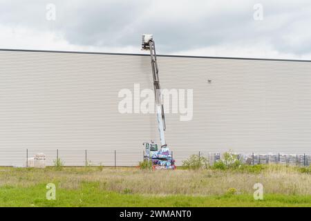 Un ouvrier soulève des matériaux sur le toit d'un entrepôt gris avec un ascenseur hydraulique. Le ciel nuageux laisse entrevoir la possibilité de pluie. Banque D'Images