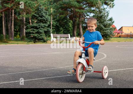 Souriant garçon mignon dans des vêtements bleus est à cheval sur un tricycle dans un parc. Banque D'Images