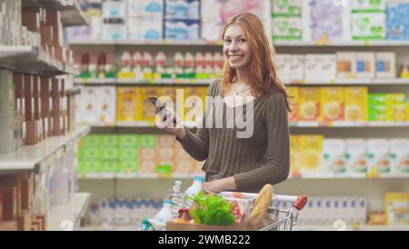 Portrait d'une jeune femme faisant l'épicerie au supermarché, elle sourit et tient son smartphone Banque D'Images