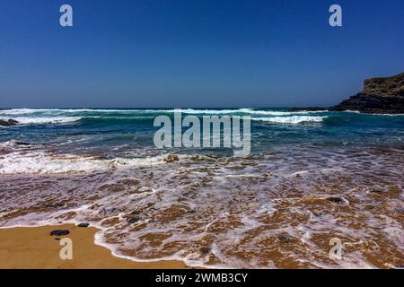 Une plage de sable avec des vagues mousseuses et des falaises rocheuses sous un ciel bleu clair. Banque D'Images