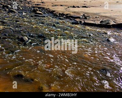 Un ruisseau coule sur des rochers, son eau brune scintillant à la lumière du soleil, entouré d'un terrain rocheux. Banque D'Images
