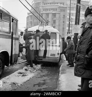 Bucarest, Roumanie, approx. 1979. Scène matinale à la gare routière en face de la gare (Gara de Nord) dans une journée d'hiver. Des hommes à l'extérieur d'un bus surpeuplé. Les gens traversent la rue à travers des flaques et de la neige fondante, la plupart d'entre eux sur le chemin du travail. Banque D'Images