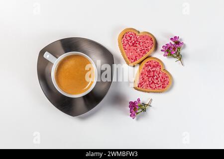 Une tasse de café sur une soucoupe avec deux biscuits en forme de coeur en glaçage rose et brins de fleurs. Vue de dessus sur fond blanc. Banque D'Images