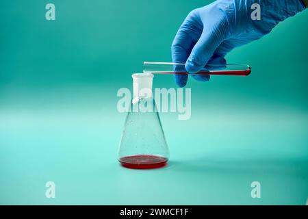 Main d'un scientifique médical anonyme dans un gant jetable versant l'échantillon liquide rouge du tube à essai dans le flacon de titrage pendant la réutilisation du laboratoire scientifique Banque D'Images