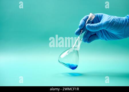 Main d'un chercheur méconnaissable dans un gant en latex tenant un flacon de titrage de substance chimique bleue pendant l'expérience en laboratoire Banque D'Images