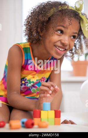 Sourire fille afro-américaine aux cheveux bouclés avec arc jaune sourires tout en empilant des blocs, robe rayée arc-en-ciel dynamique. pour le contenu sur la joie dans l'apprentissage Banque D'Images