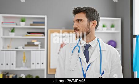 Beau jeune homme hispanique en blouse de laboratoire blanche avec stéthoscope, regardant réfléchi à la clinique. Banque D'Images