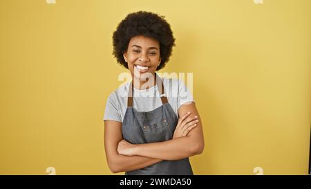 Portrait d'une femme souriante aux cheveux bouclés et aux bras croisés portant un tablier sur un fond jaune. Banque D'Images