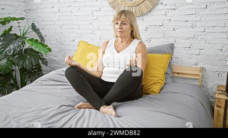 Éveillée et concentrée, femme blonde d'âge moyen aime l'exercice de yoga matinal assis sur le lit, embrassant le calme et l'équilibre dans sa chambre à coucher Banque D'Images
