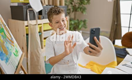 Adorable garçon artiste blond, souriant lors d'un appel vidéo depuis son studio d'art, toile et pinceau à portée de main, entièrement immergé dans son apprentissage créatif jou Banque D'Images