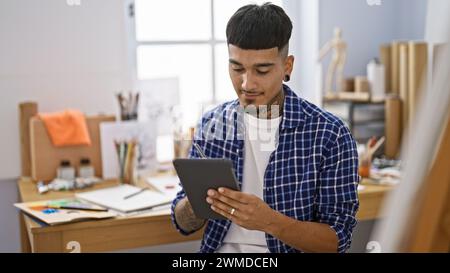 Tatoué jeune homme latin dessinant joyeusement sur le touchpad, peaufinant la créativité numériquement au studio d'art. Banque D'Images