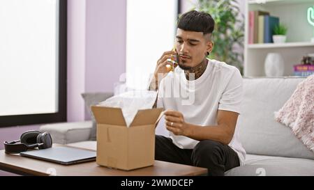 Tatoué homme latin parlant intentionnellement au téléphone tout en déballant la boîte de livraison dans son appartement de salon Banque D'Images