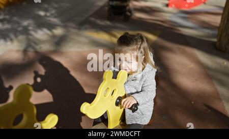 Jolie fille blonde jouant sur un cavalier de printemps en forme d'animal jaune de terrain de jeu dans un parc ensoleillé. Banque D'Images