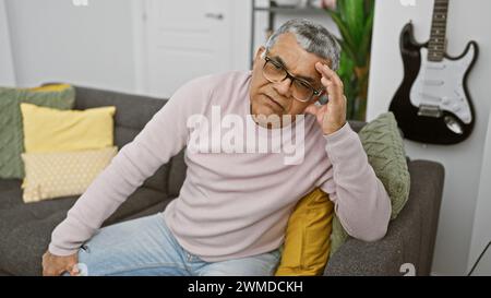 Portrait d'un homme senior pensif avec des lunettes, assis sur un canapé à l'intérieur avec une guitare dans le fond flou. Banque D'Images