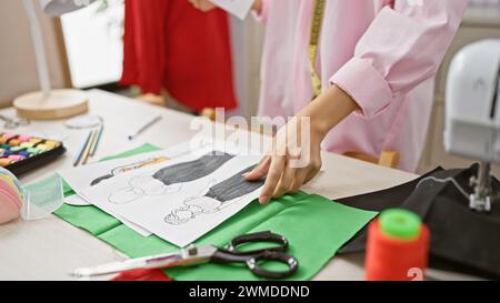 Un gros plan des mains d'une créatrice de mode féminine travaillant avec du tissu et des croquis sur un bureau dans un atelier. Banque D'Images