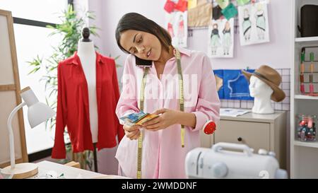 Une jeune femme hispanique compte des francs suisses dans son magasin de vêtements urbains, entourée de matériel de couture et de créations de mode. Banque D'Images