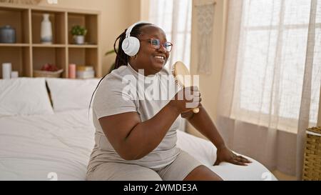 Femme afro-américaine confiante appréciant le matin, se relaxant sur le lit chantant à une chanson en utilisant une brosse comme microphone, entendant sa voix à travers la tête Banque D'Images
