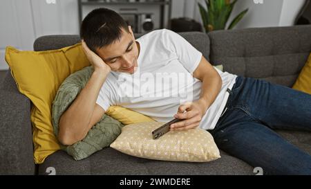 Un jeune homme hispanique détendu utilise tranquillement un smartphone tout en se prélassant sur un canapé confortable avec des oreillers décoratifs dans un salon moderne. Banque D'Images