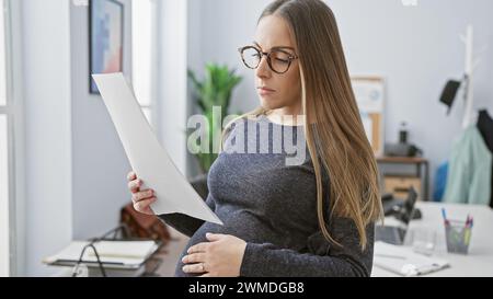 Une femme hispanique enceinte lit un document dans un bureau, décrivant la maternité sur le lieu de travail. Banque D'Images
