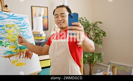 Jeune artiste chinois confiant, portant un tablier d'abricot, dessine et peint avec joie à l'intérieur dans un studio d'art universitaire tout en souriant pendant un vid animé Banque D'Images