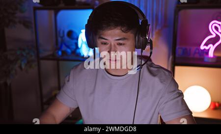 Un jeune homme asiatique portant des écouteurs se concentre intensément dans une salle de jeu éclairée d'ambiance la nuit. Banque D'Images