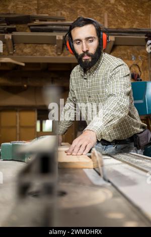 Le menuisier vertical avec protection auditive utilise une scie à table pour couper une planche de bois dans un atelier de menuiserie bien équipé. Conc. Business Banque D'Images