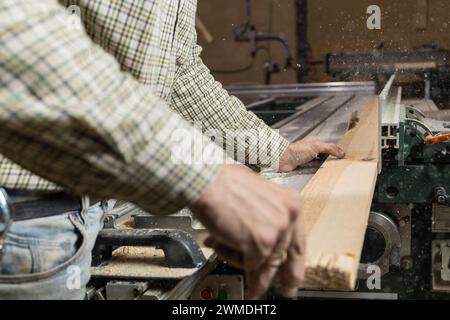 Photo horizontale vue rapprochée des mains d'un charpentier alors qu'il introduit manuellement un morceau de bois dans une scie à table, avec de la sciure tourbillonnant dans l'atelier. Bu Banque D'Images