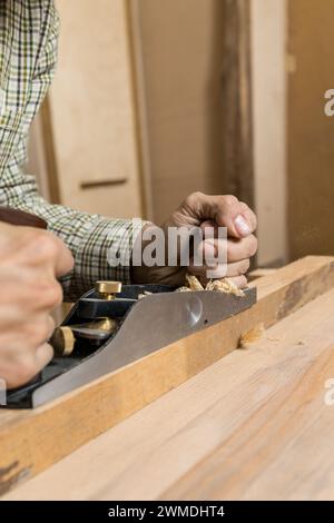 Photo verticale vue détaillée des mains d'un charpentier comme ils utilisent habilement un plan de main pour lisser une planche de bois, avec des copeaux de bois volant. Copier Banque D'Images