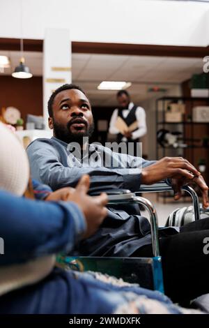 Jeune homme afro-américain en fauteuil roulant arrivant à l'hôtel de luxe avec des installations pour handicapés, homme handicapé voyageant avec sa femme. Tourisme accessible, hébergement de voyage et handicap Banque D'Images