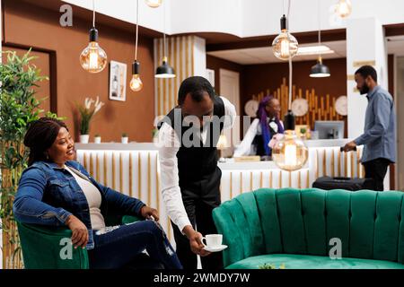 Heureuse femme afro-américaine souriante touriste se détendant dans le hall de l'hôtel, regardant le serveur servant le café, se sentant satisfaite du service à la clientèle. Amical employé de restaurant mettant la tasse sur la table Banque D'Images