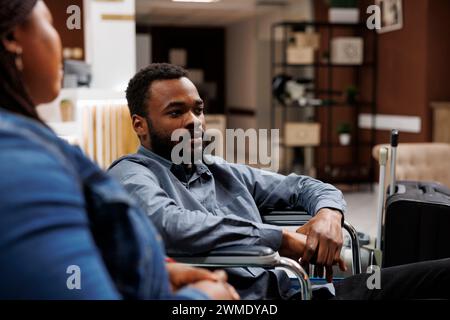 Homme afro-américain touriste avec handicap physique en fauteuil roulant attendant avec bagages dans le hall de l'hôtel, arrivant au centre de réadaptation pour handicapés. Tourisme accessible inclusif Banque D'Images