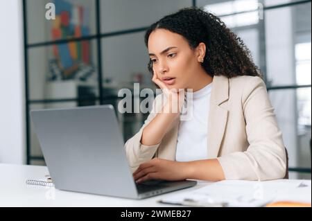 Bouleversée brésilienne ou hispanique aux cheveux frisés, chef de la direction de l'entreprise, directeur financier, siège dans un bureau créatif moderne, regarde tristement un ordinateur portable, étudie l'information, lit de mauvaises nouvelles Banque D'Images