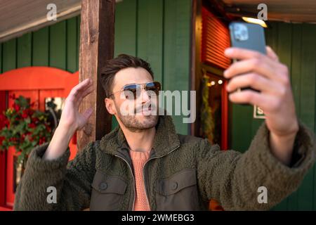 Beau jeune homme en veste tendance et lunettes de soleil prenant selfie avec smartphone. Hipster masculin faisant chat vidéo via téléphone portable à l'extérieur de la maison en bois. Banque D'Images