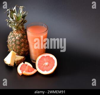 Un mélange de jus de fruits tropicaux fraîchement pressés sur fond noir, à côté des moitiés de pamplemousse mûr et d'ananas. Gros plan. Banque D'Images