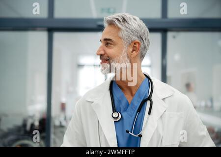 Portrait d'un médecin adulte confiant debout dans le couloir de l'hôpital. Beau médecin avec les cheveux gris portant un manteau blanc, gommages, stéthoscope autour du cou Banque D'Images