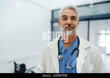 Portrait d'un médecin adulte confiant debout dans le couloir de l'hôpital. Beau médecin avec les cheveux gris portant un manteau blanc, gommages, stéthoscope autour du cou Banque D'Images