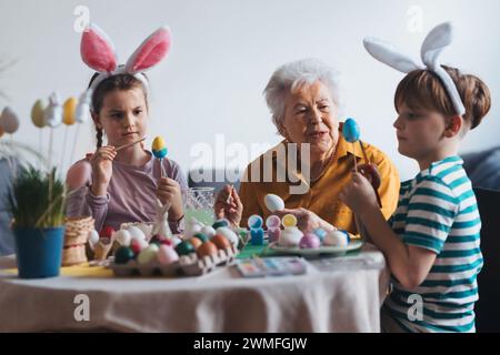 Grand-mère avec de petits enfants décorant des œufs de pâques à la maison. Tradition de peindre les oeufs avec pinceau et colorant d'oeuf de pâques. Banque D'Images