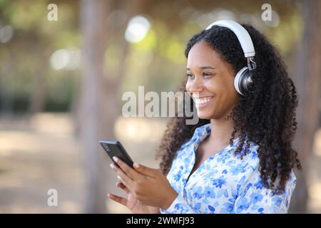 Femme noire heureuse portant casque écoutant de la musique tenant le téléphone à l'extérieur Banque D'Images