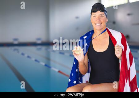 Une femme caucasienne présente sa médaille d'or au bord de la piscine avec un drapeau américain Banque D'Images