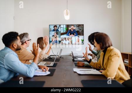 Une équipe diversifiée de professionnels participe avec enthousiasme à une visioconférence, agitant la main, reflétant une culture d'entreprise chaleureuse et inclusive qui embrasse la collaboration à distance et l'unité d'équipe Banque D'Images