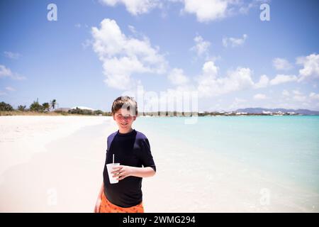 Tween Boy Smiles à Camera, tenant un verre sur Caribbean Beach Banque D'Images