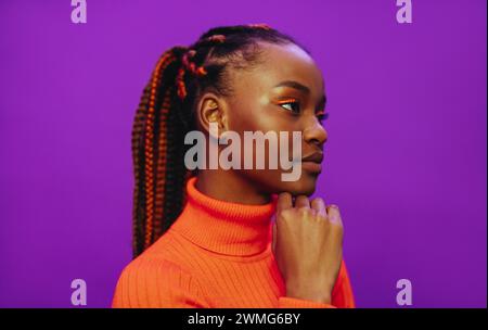 Confiant gen Z femme dans sa vingtaine avec des tresses élégantes, un maquillage vibrant et des vêtements décontractés pose sur un fond violet avec portrait en gros plan. Banque D'Images