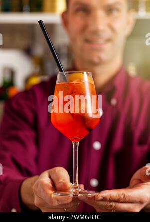 Barman souriant dans une chemise violette vibrante servant un spritz aperol rafraîchissant dans un bar Banque D'Images