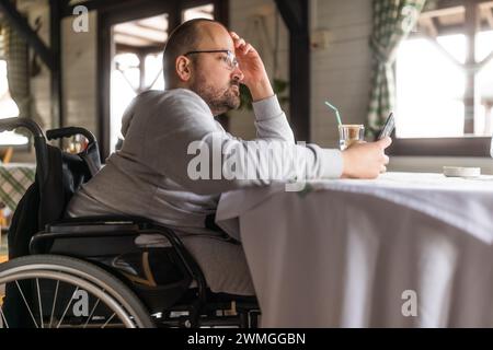 Homme handicapé paraplégique en fauteuil roulant est assis au restaurant et utilise un smartphone. Banque D'Images