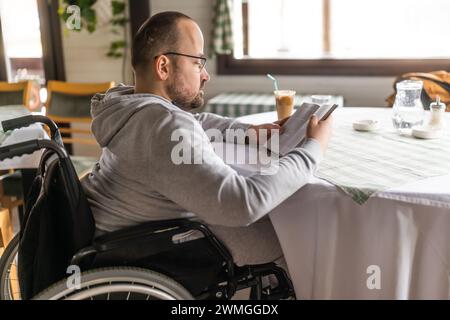 L'homme handicapé paraplégique en fauteuil roulant est assis au restaurant et lit un livre. Banque D'Images